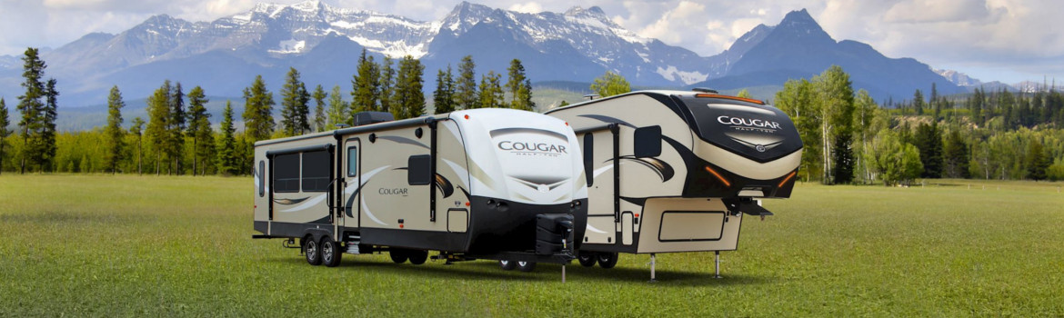 A Cougar fifth wheel and travel trailer side by side in an open field next to mountains